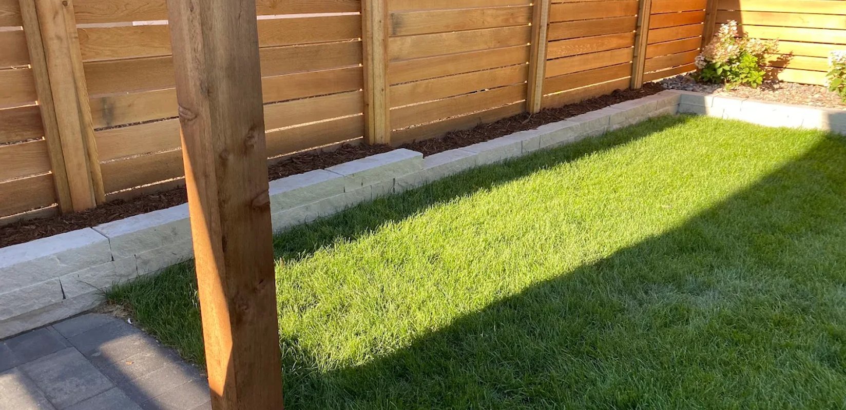 A newly installed structural retaining wall and pressure-treated wood fence built in an Edmonton residential backyard to manage a steep slope and provide privacy.